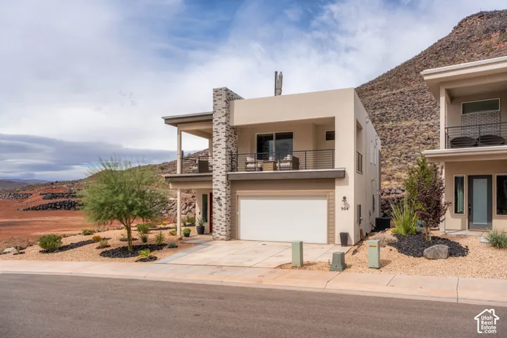 Modern exterior featuring a stucco facade, stone-accented chimney, concrete driveway, and a spacious covered balcony with metal railing
