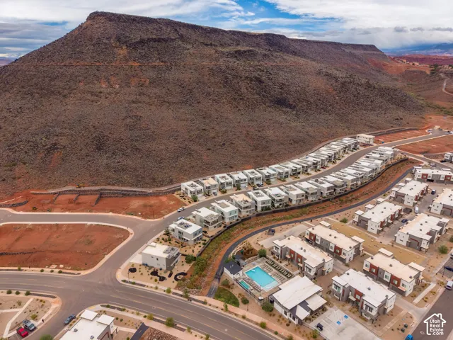 Aerial perspective featuring a multi-unit residential community situated against a prominent mountain backdrop, inclusive of an on-site swimming pool, paved roadways, and desert landscaping
