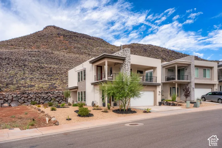 Modern two-story exterior featuring a stone-clad facade, stucco finish, attached garage, and a second-level balcony with metal railing, set against a prominent mountain backdrop