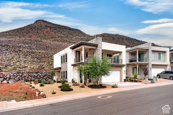 Contemporary two-story exterior featuring smooth stucco siding, stone masonry accents, a covered second-story balcony with metal railings, and a multi-car garage, set against a backdrop of natural mountain terrain