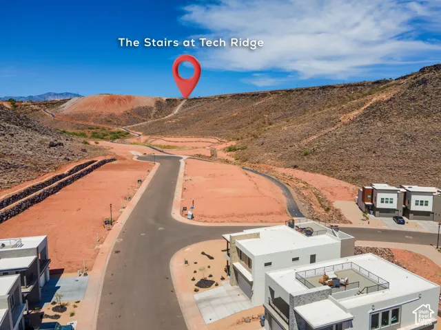 Aerial perspective of a residential development featuring paved streets, multi-level modern architecture with flat roofing, rooftop terraces, and bordering desert hillside terrain