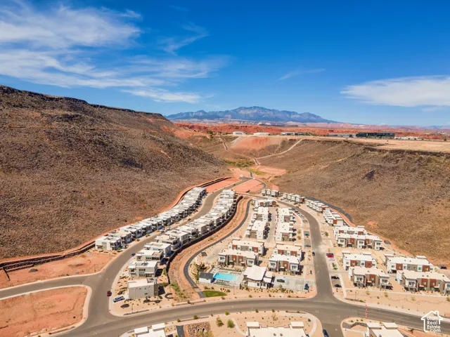 Aerial perspective of a residential development featuring multi-level dwellings, paved roadways, and a communal pool, set against a backdrop of desert terrain and mountain ranges