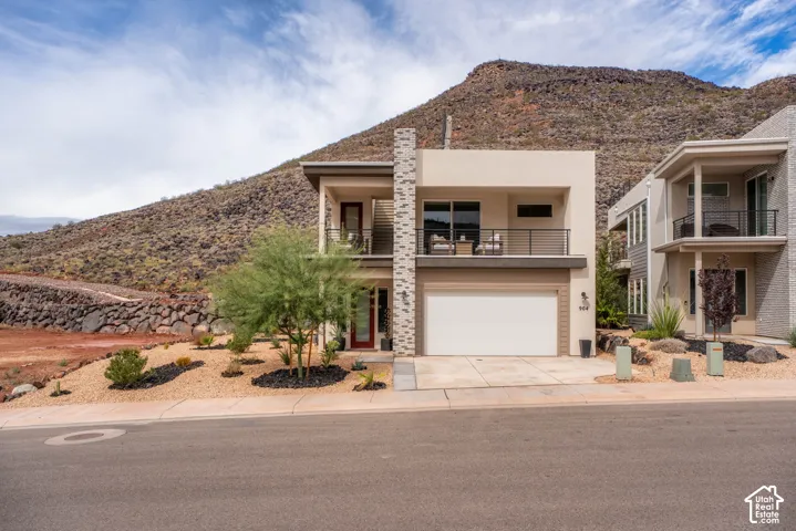Contemporary residence featuring a stone-clad chimney, stucco exterior, upper-level balcony with metal railing, two-car garage, and desert-inspired xeriscaping
