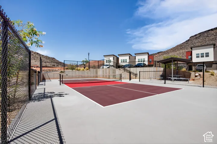 Pickleball court with a multi-toned surface, enclosed by chain-link fencing, accompanied by a shaded seating area and concrete retaining walls