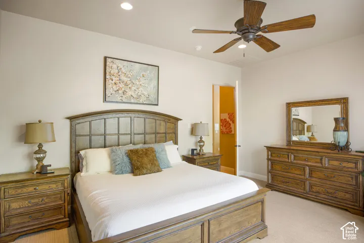 Spacious primary bedroom featuring a wood-finish bed frame with metal headboard detailing, wood-finish nightstands with table lamps, a wood-finish dresser with mirror, ceiling fan with light fixture, recessed lighting, and neutral wall-to-wall carpeting t