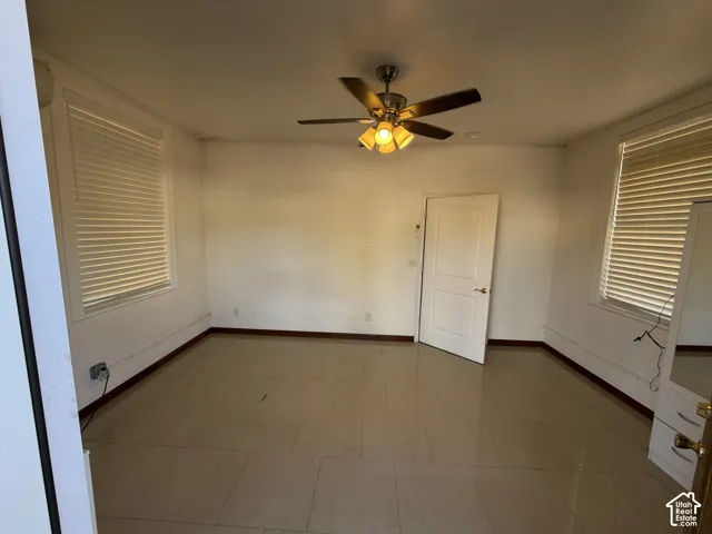 Neutral-toned tile flooring, dual windows with fitted horizontal blinds, ceiling-mounted fan with integrated light fixture, and a solid-core interior door