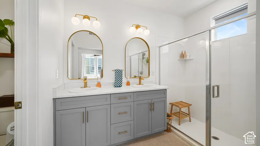 Double vanity featuring gray cabinetry, brass-toned hardware, arched mirrors with gold-tone frames, and a glass-enclosed shower with a built-in seat
