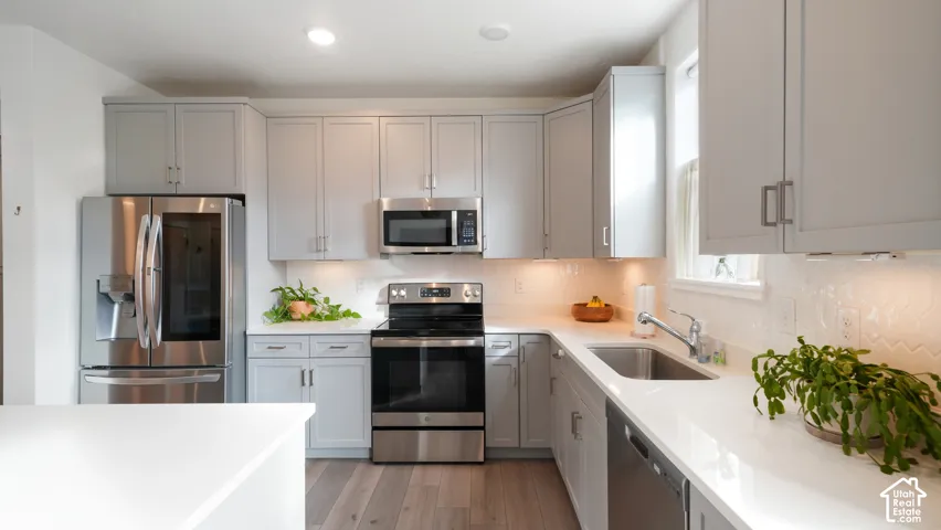 Kitchen featuring light-gray Shaker-style cabinetry, white quartz countertops, herringbone-patterned tile backsplash, stainless steel appliances, and wood-finish flooring
