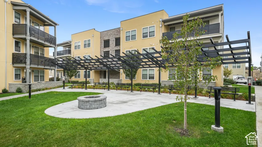 Outdoor common area featuring a circular stone fire pit, extensive metal pergola, concrete hardscaping, manicured lawn, and perimeter metal benches