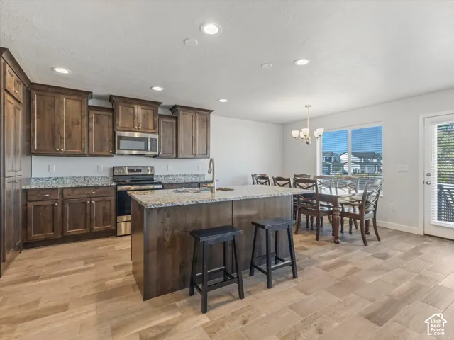 Kitchen featuring stained wood cabinetry, granite countertops, a central island with an integrated sink, stainless steel appliances, and wood-finish tile flooring