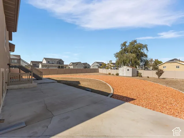 Expansive backyard featuring a concrete patio, wood-toned elevated deck, decorative stone landscaping, and a detached storage shed, all enclosed by vinyl perimeter fencing