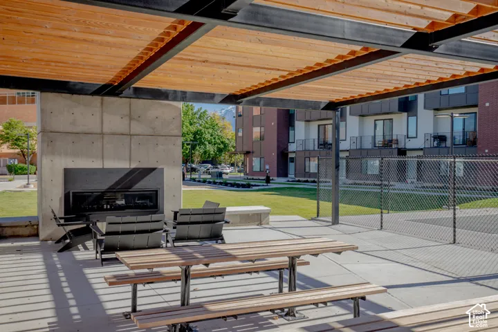 Covered outdoor pavilion featuring a timber-slat pergola, black steel support beams, concrete fireplace, and built-in wood-slat picnic seating