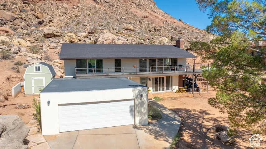 Two-story residence featuring an upper-level balcony with metal railing, a detached storage structure, concrete driveway, and a natural rock formation backdrop