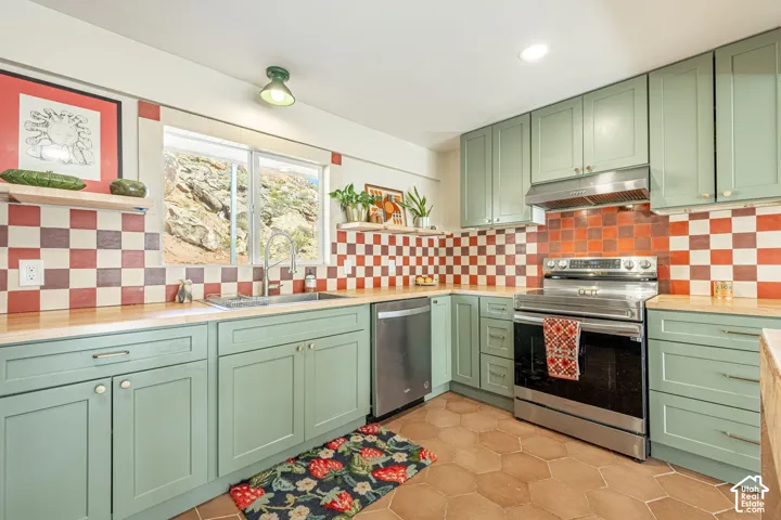 Kitchen featuring sage green cabinetry, butcher block countertops, stainless steel appliances, checkered tile backsplash, and hexagonal terra-cotta tile flooring