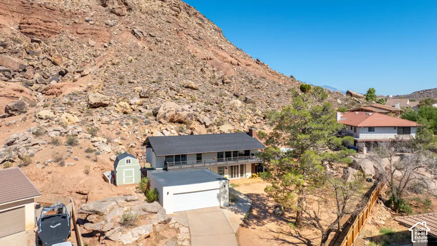 Two-story residence featuring an attached garage, upper-level balcony, shingled roof, and natural rocky hillside terrain