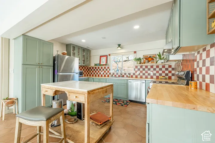 Kitchen featuring sage green Shaker-style cabinetry, butcher block countertops, a checkerboard tile backsplash, and hexagonal terra cotta floor tiles