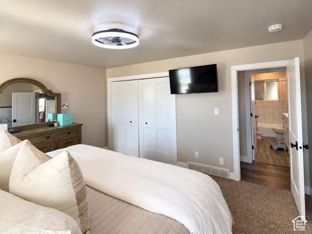 Neutral-toned room featuring a contemporary circular LED ceiling fan, double-panel closet doors with matte black hardware, and an open doorway revealing a tiled bathroom with wood-finish flooring