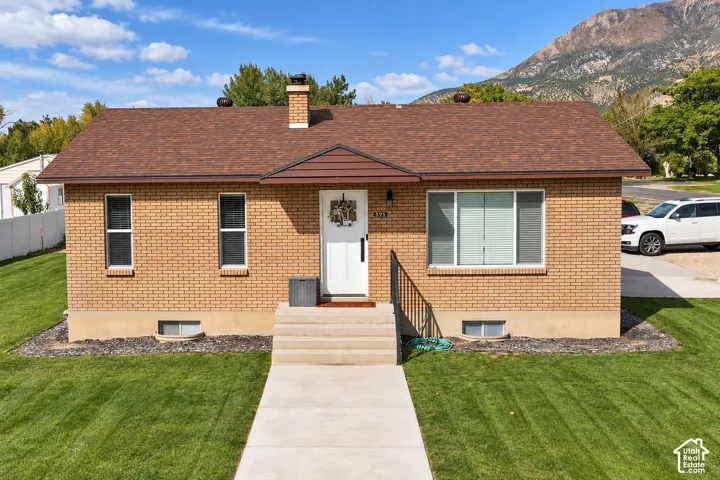 Single-level brick exterior featuring a shingle roof, concrete entryway with metal handrails, mountain backdrop, and expansive lawn