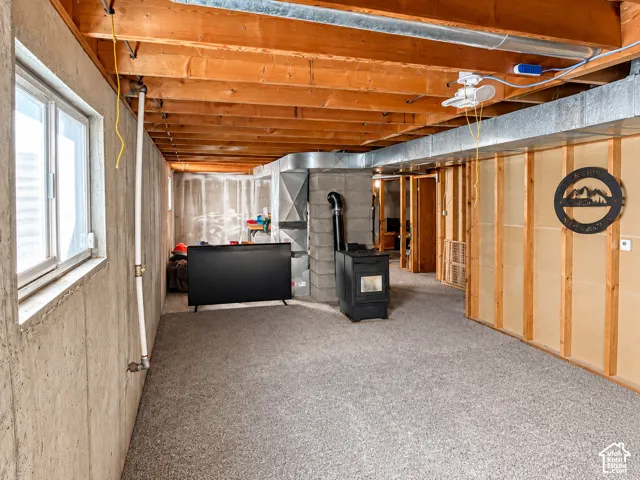 Unfinished interior space featuring exposed wood joist ceilings, concrete wall construction, grey carpeting, and a metal pellet stove with integrated flue