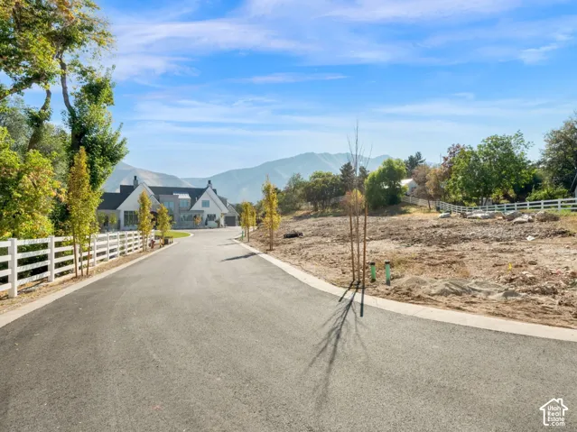 Paved driveway with a white split-rail fence, stone pillar, and distant mountain backdrop