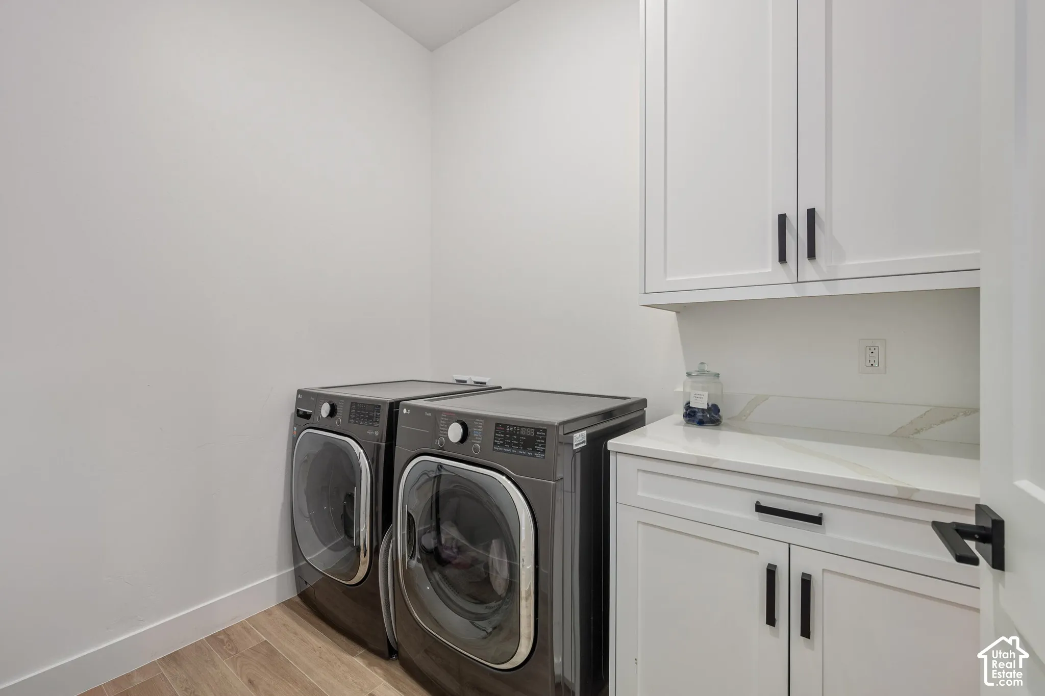 Adjacent to Primary bedroom Laundry space featuring white shaker-style cabinetry, stone-surface countertop, wood-finish flooring, and matte black hardware