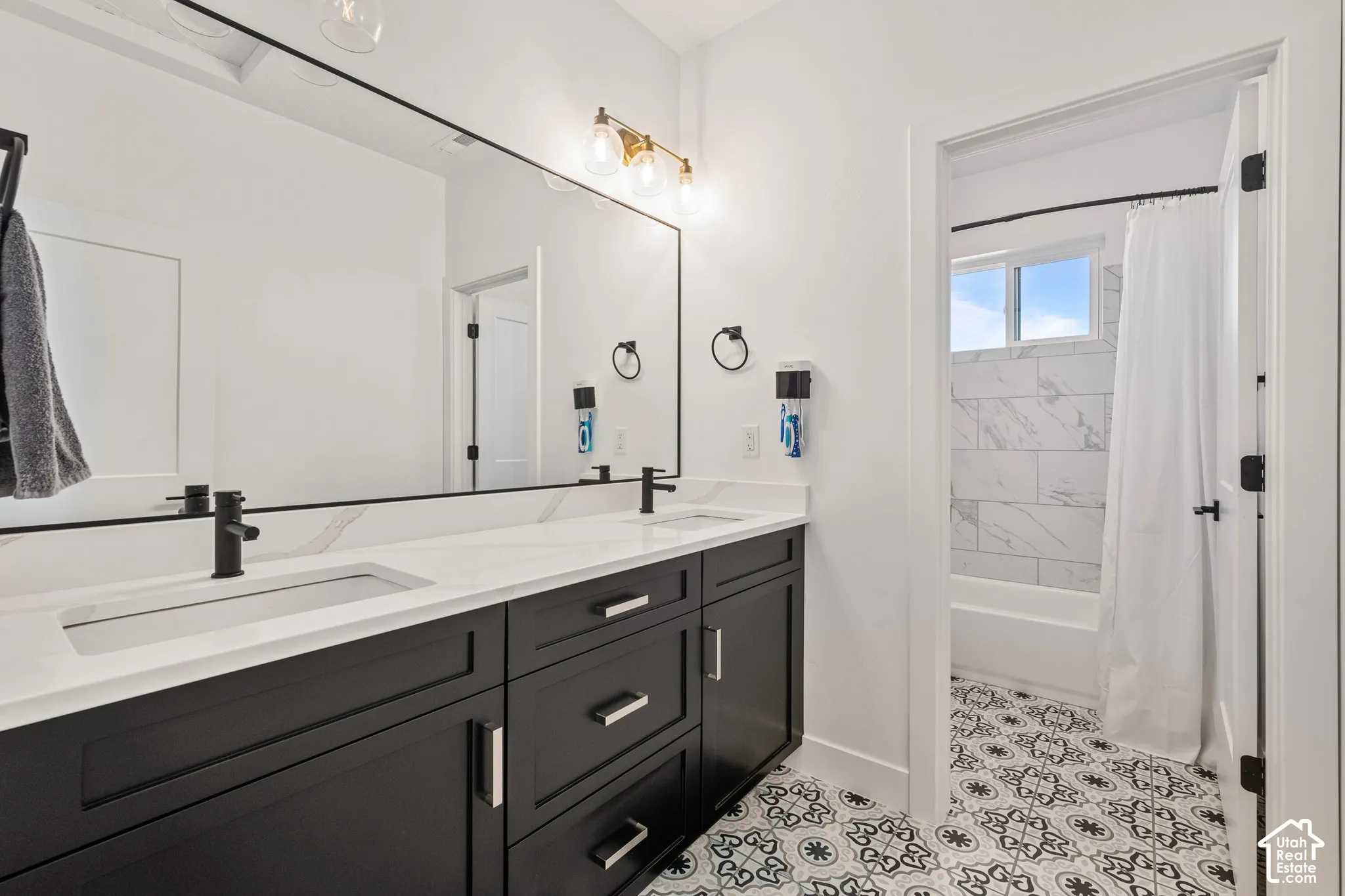 Bathroom featuring a dual-sink vanity with a stone countertop, matte black faucets, patterned tile flooring, and a tub-shower combination with marble-finish wall tile