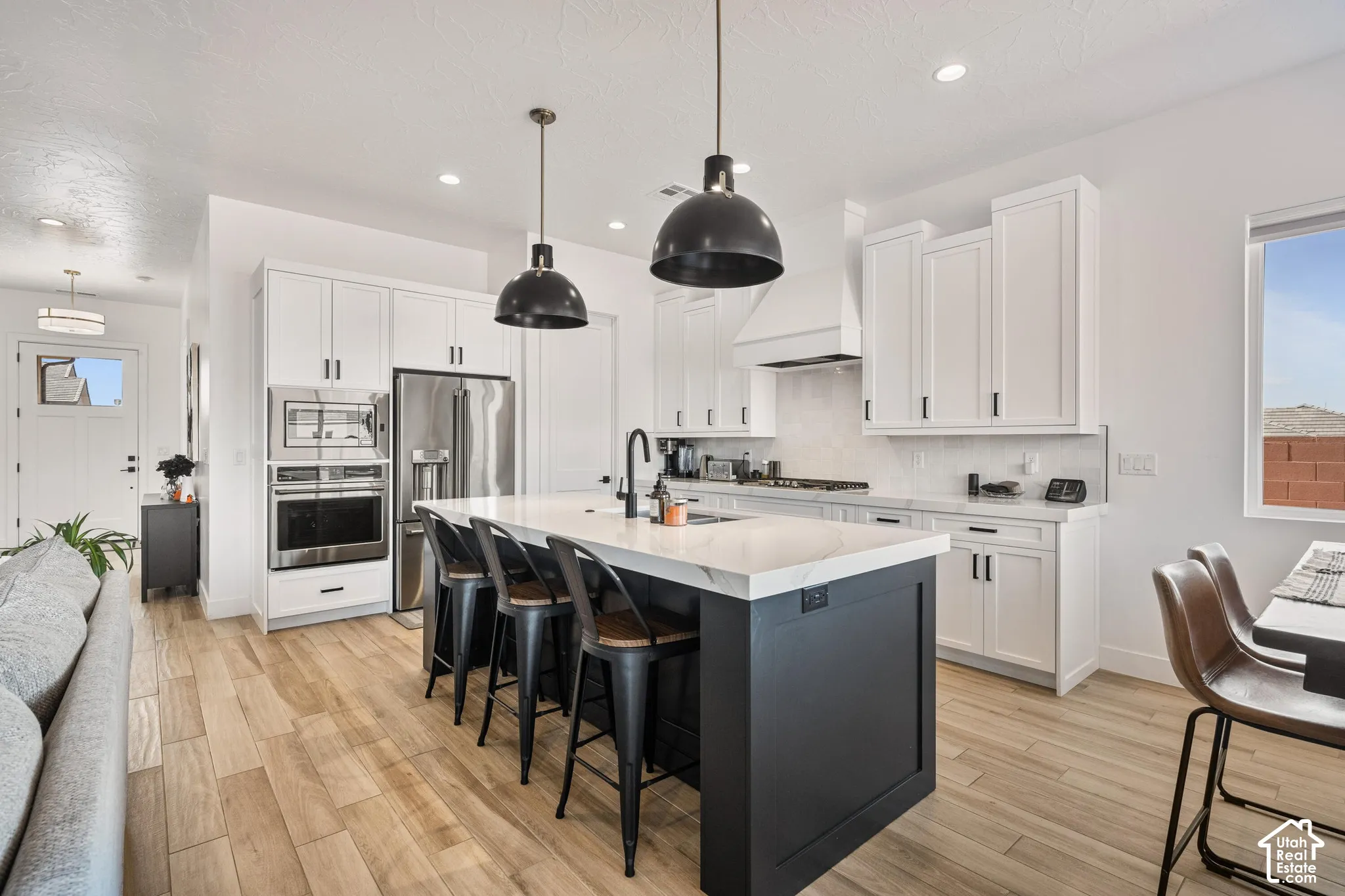 Kitchen featuring a dark-toned center island with a stone countertop, white shaker-style cabinetry, stainless steel built-in appliances, pendant lighting, and wood-finish flooring