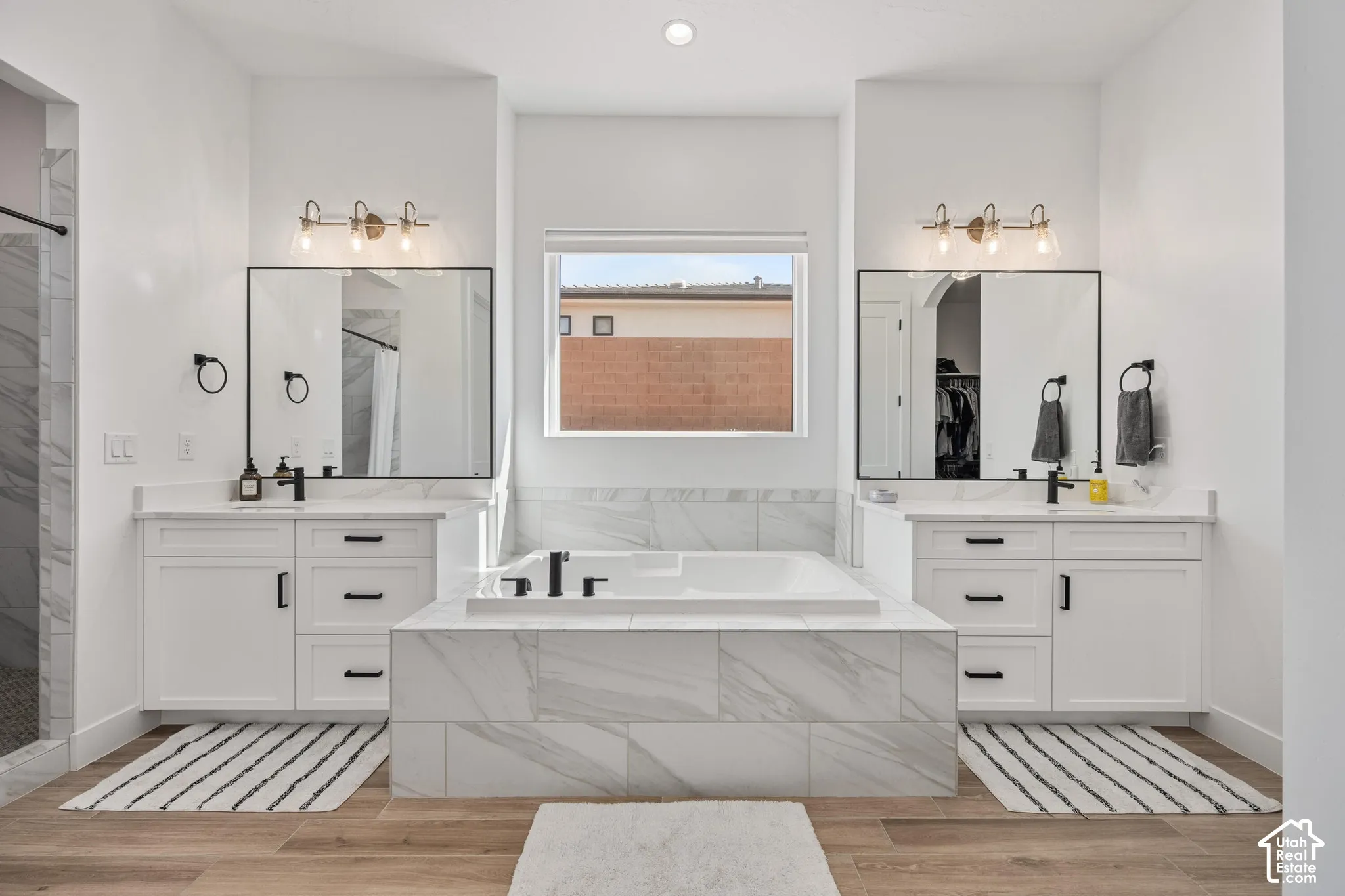 Primary bath Dual vanities with white shaker-style cabinetry, rectangular undermount sinks, matte black fixtures, and stone-finish countertops, centered around a tiled soaking tub and wood-finish flooring
