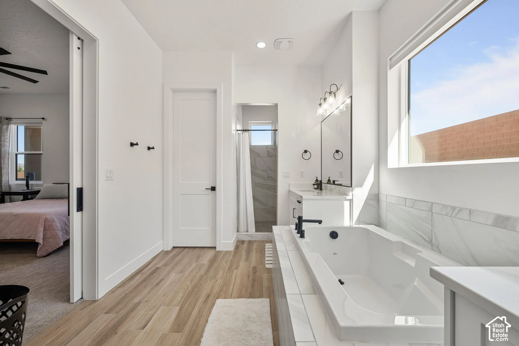 Primary Bathroom featuring a deep soaking tub with tile surround, wood-finish flooring, white shaker-style cabinetry, matte black plumbing fixtures, and a large picture window