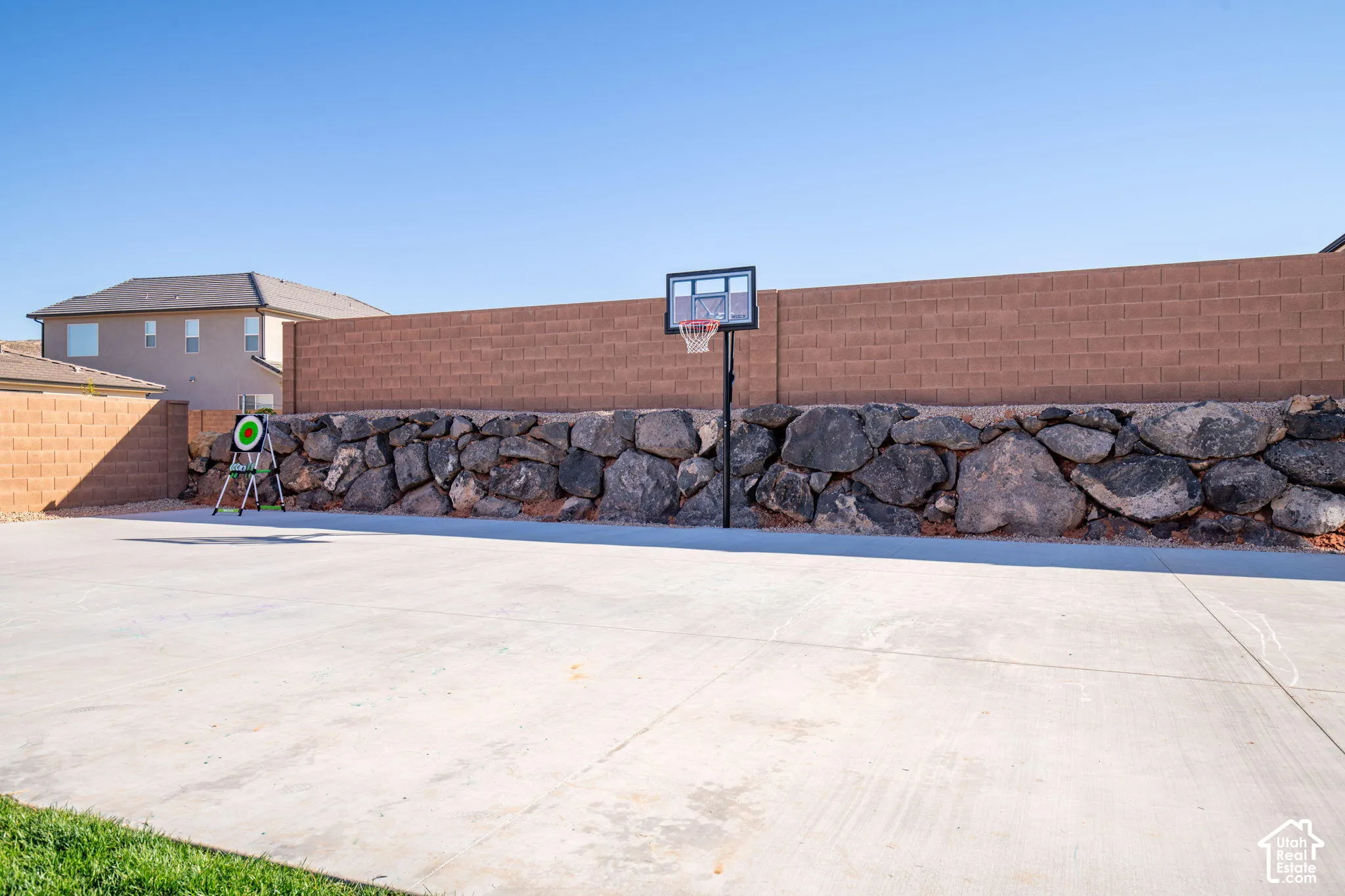 Expansive concrete pickleball sized sport court featuring a masonry block wall, built-in basketball hoop, and natural stone retaining wall