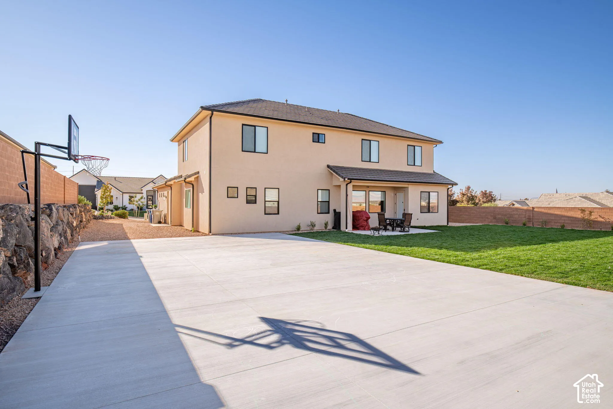 Two-story stucco exterior featuring a large concrete patio, manicured lawn, stone retaining wall, and an integrated basketball standard