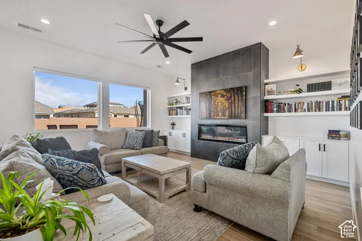 Living area featuring a floor-to-ceiling tile fireplace surround, linear electric insert, built-in shelving with cabinetry, wood-finish flooring, and a multi-blade ceiling fan
