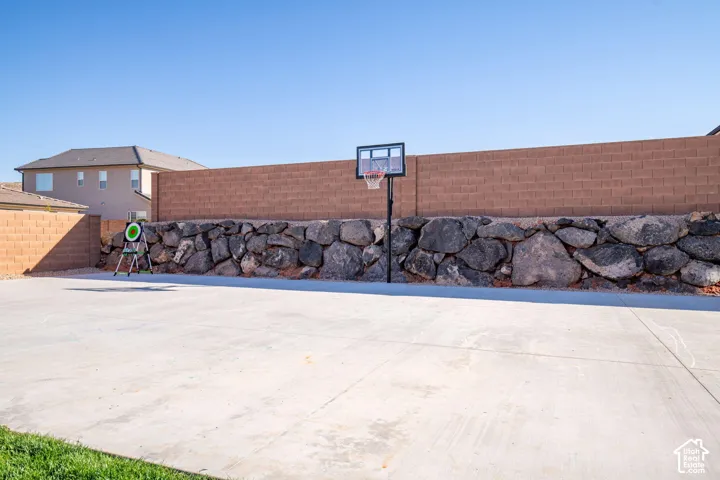 Expansive concrete pickleball sized sport court featuring a masonry block wall, built-in basketball hoop, and natural stone retaining wall