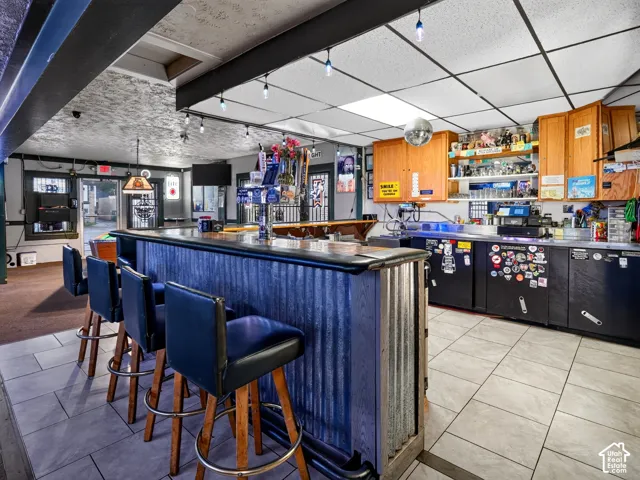 Bar area featuring a corrugated metal front, wood-finish cabinetry, tile flooring, drop ceiling with integrated lighting, and a built-in service counter