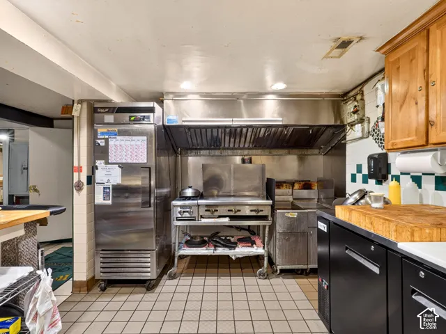 Commercial-grade kitchen featuring a stainless steel reach-in refrigerator, heavy-duty stainless steel range with overhead ventilation hood, tiled flooring, and wood-finish cabinetry