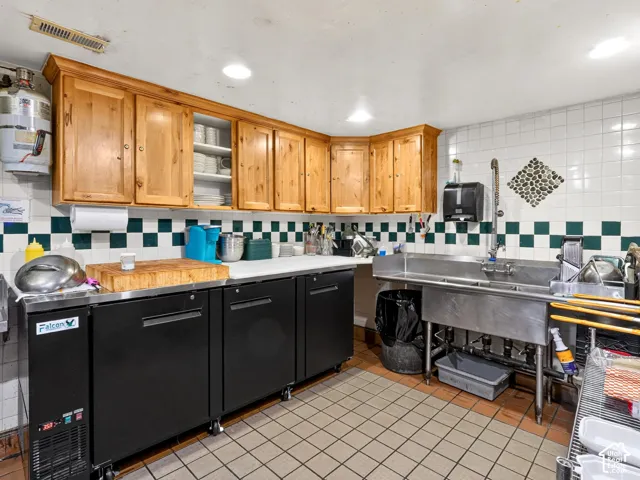 Commercial-grade kitchen featuring stainless steel triple-basin sink with pull-down sprayer, wood cabinetry, stone-tiled backsplash, and built-in black under-counter refrigeration