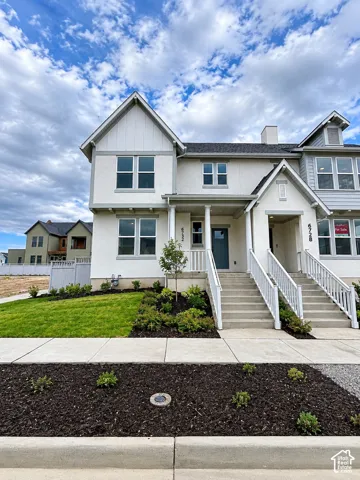 Two-story exterior featuring light-toned stucco, board and batten siding, gable rooflines, white picket-style railings, and a landscaped front yard with mulched garden beds
