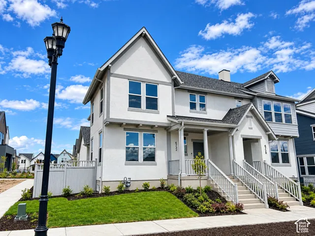 Two-story exterior featuring a light-toned stucco finish with grey trim, a covered front porch with columns, a pitched roofline, and manicured landscaping