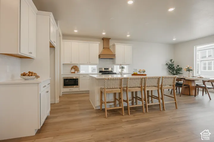 Kitchen featuring white shaker-style cabinetry, a quartz-topped center island, wood-finish flooring, a stainless steel built-in microwave, and a custom-style range hood
