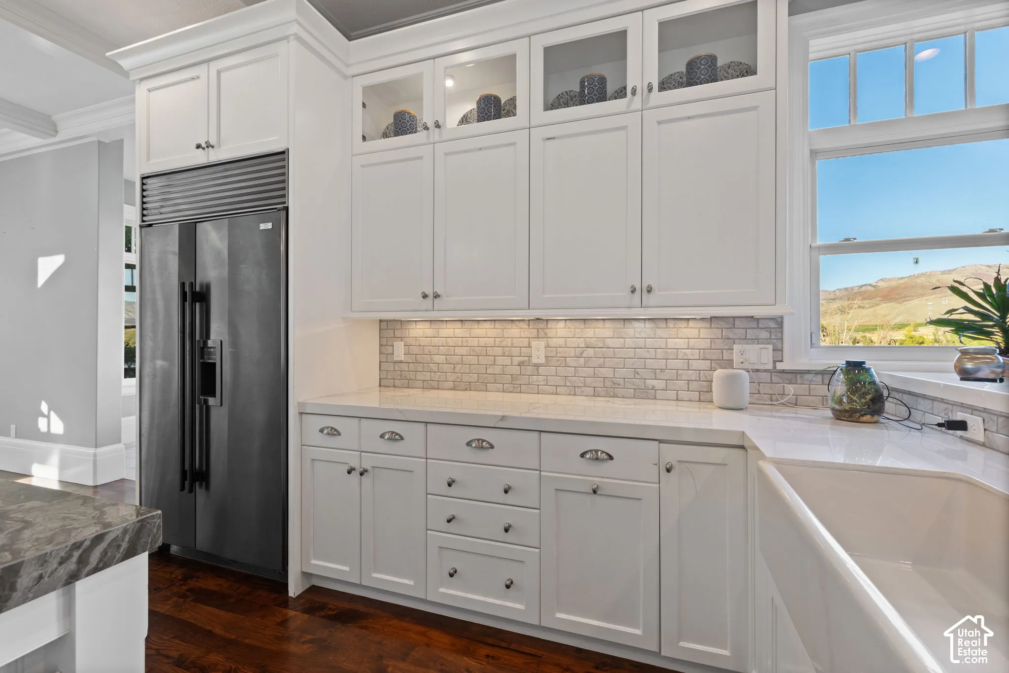 Kitchen featuring white cabinetry with glass-front uppers, solid surface countertops, a subway tile backsplash, farmhouse sink, and stainless steel integrated refrigerator