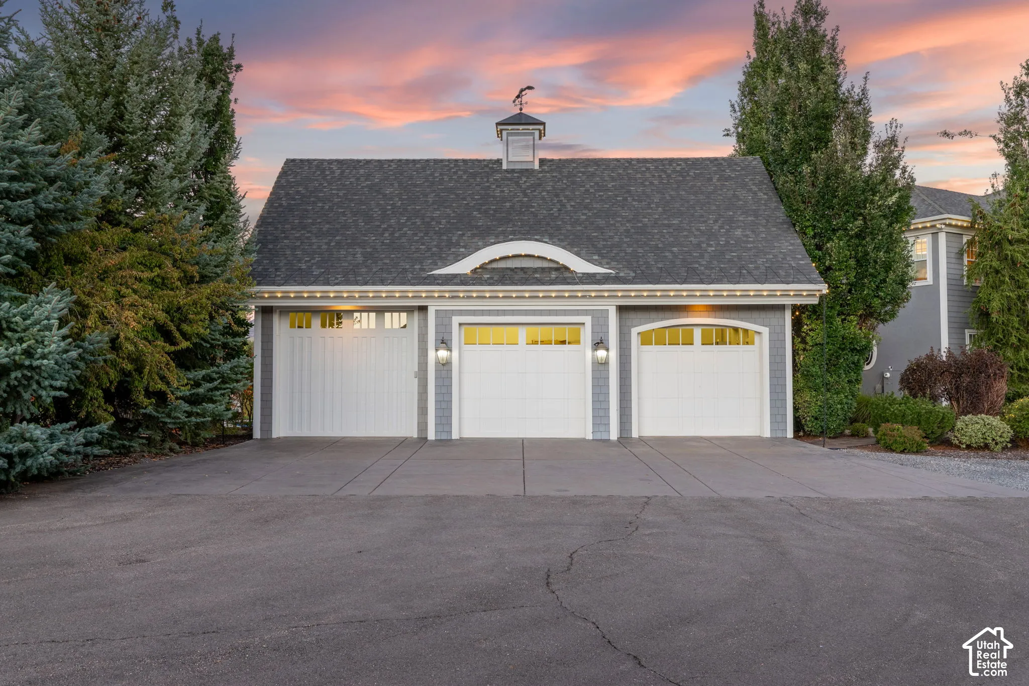 Three-car garage featuring shingle siding, an asphalt shingle roof with a cupola, arched transom windows, and exterior wall-mounted carriage lanterns