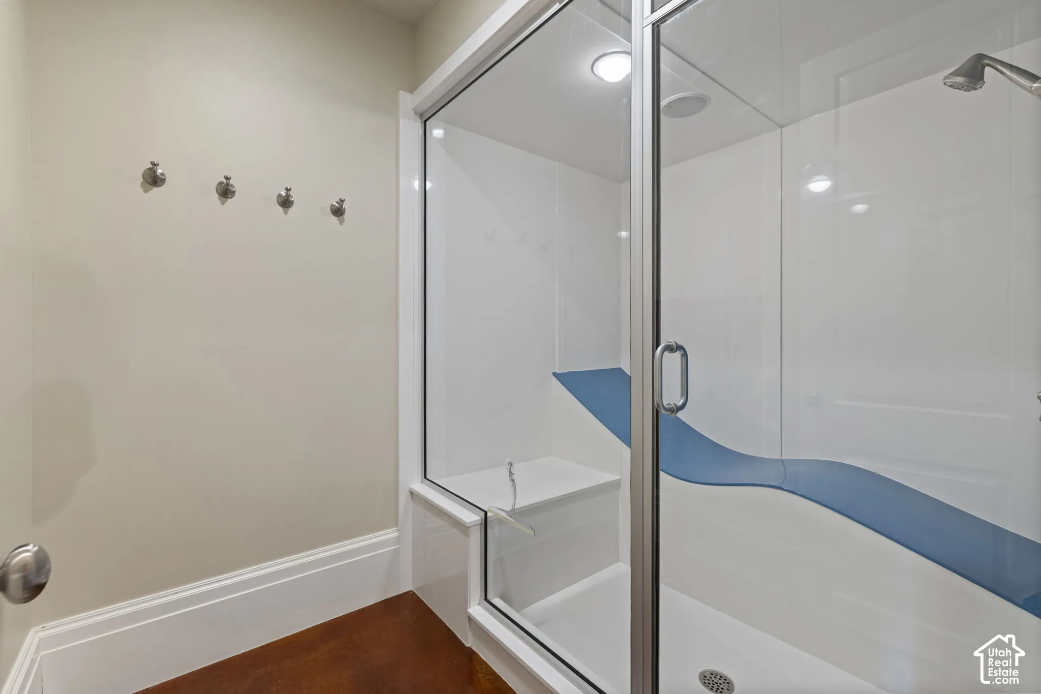 Steam shower enclosure featuring glass doors, white surround panels, an integrated bench seat, and a wall-mounted shower head, finished with wood-finish flooring and four brushed metal wall-mounted hooks