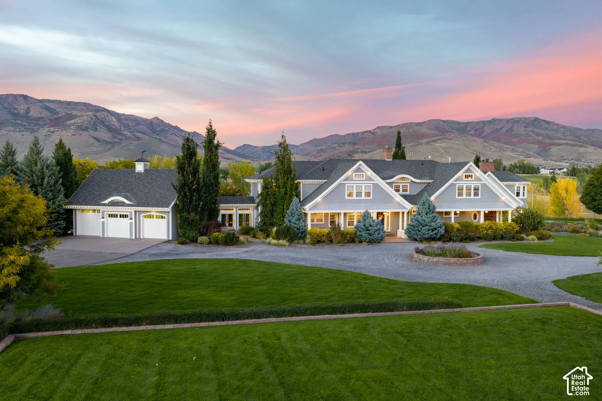 Lakefront estate featuring a multi-gabled roofline, three-car garage, circular gravel driveway, manicured lawn, and expansive mountain backdrop
