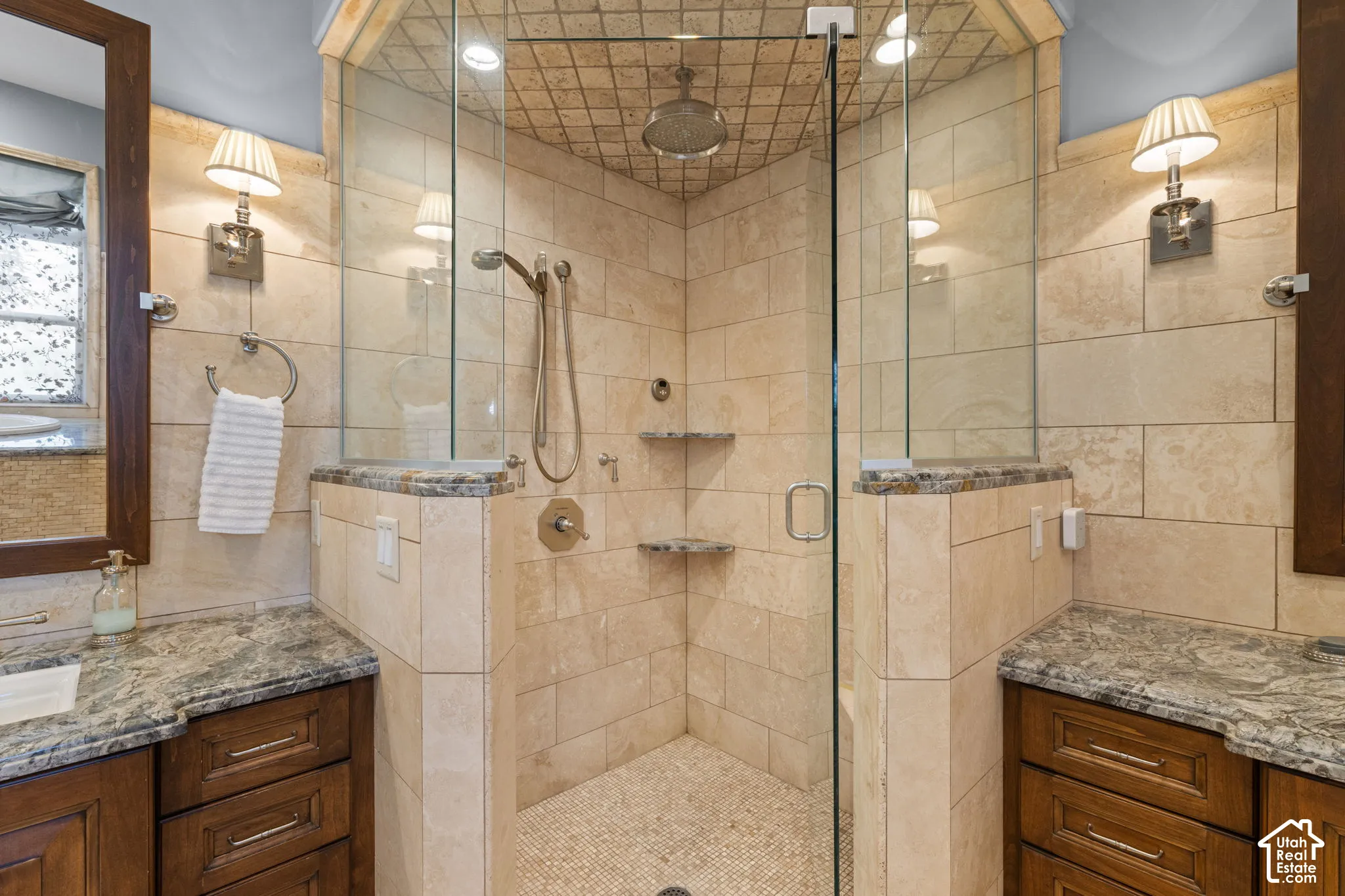 Walk-in shower with stone tile surround, integrated shelving, rainfall showerhead, and glass enclosure, flanked by dual vanities with stone countertops and wood-finish cabinetry
