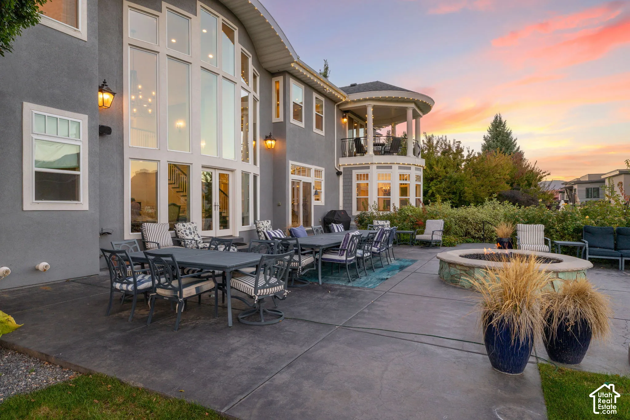 Expansive concrete patio featuring a stone-clad circular fire pit, multi-story floor-to-ceiling windows, second-story covered balcony, and exterior wall-mounted lighting