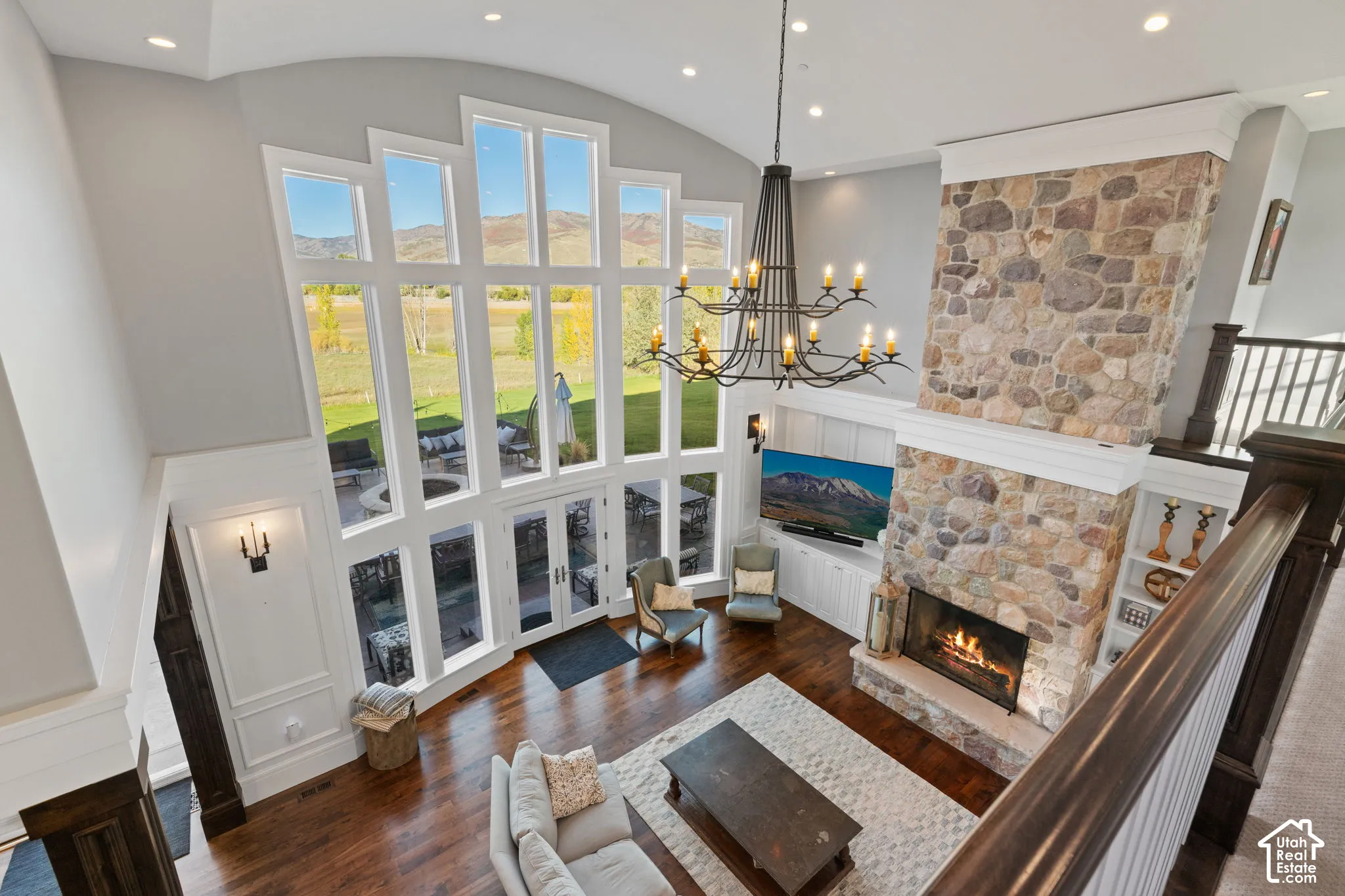 Double-height great room featuring a floor-to-ceiling multi-pane window wall, full-height stone fireplace, wood-finish flooring, custom built-in cabinetry, and a tiered iron chandelier