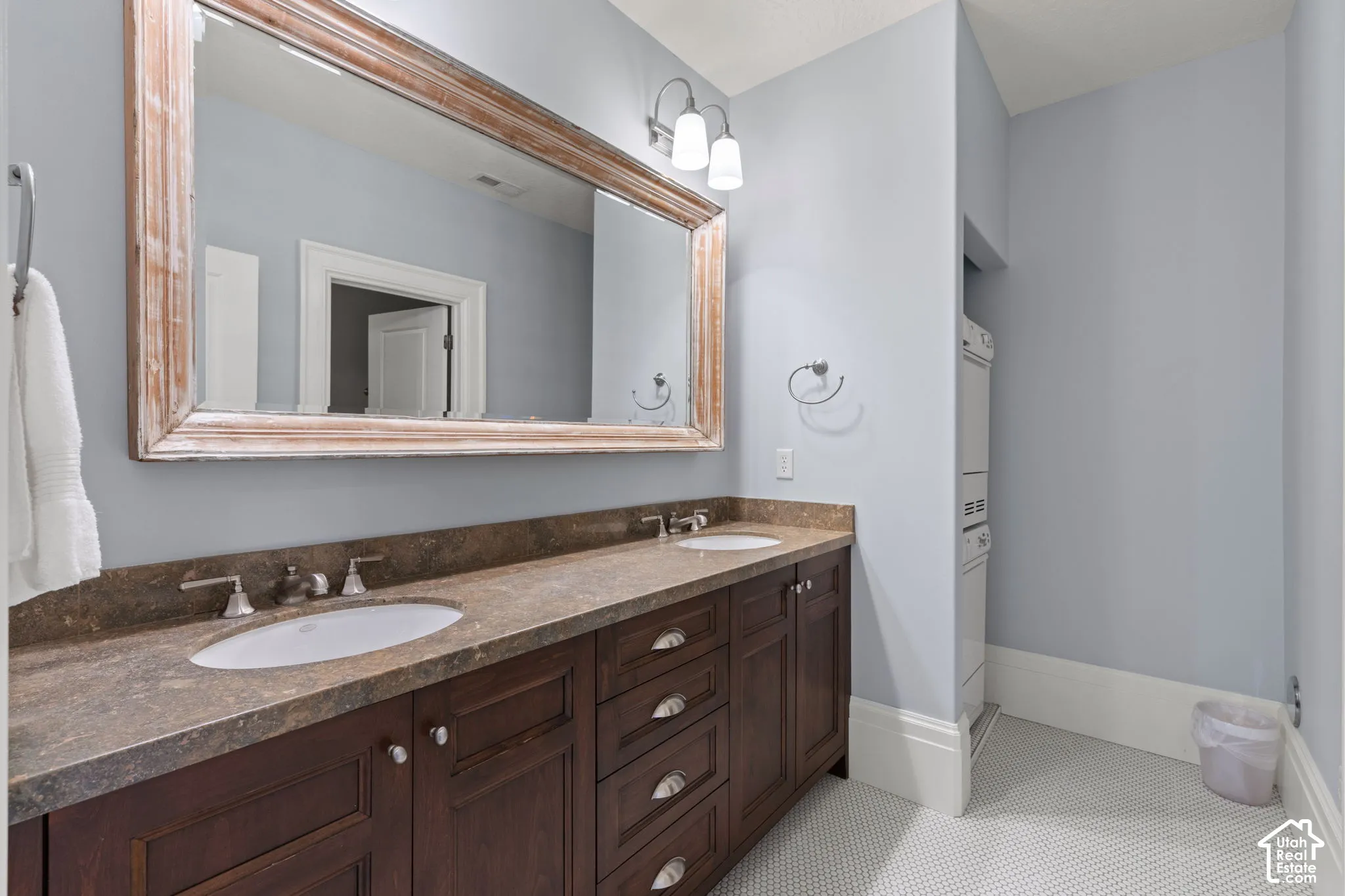 Dual-sink vanity with dark wood cabinetry, stone-finish countertops, wall-mounted light fixture, and large framed mirror, stacked washer and dryer