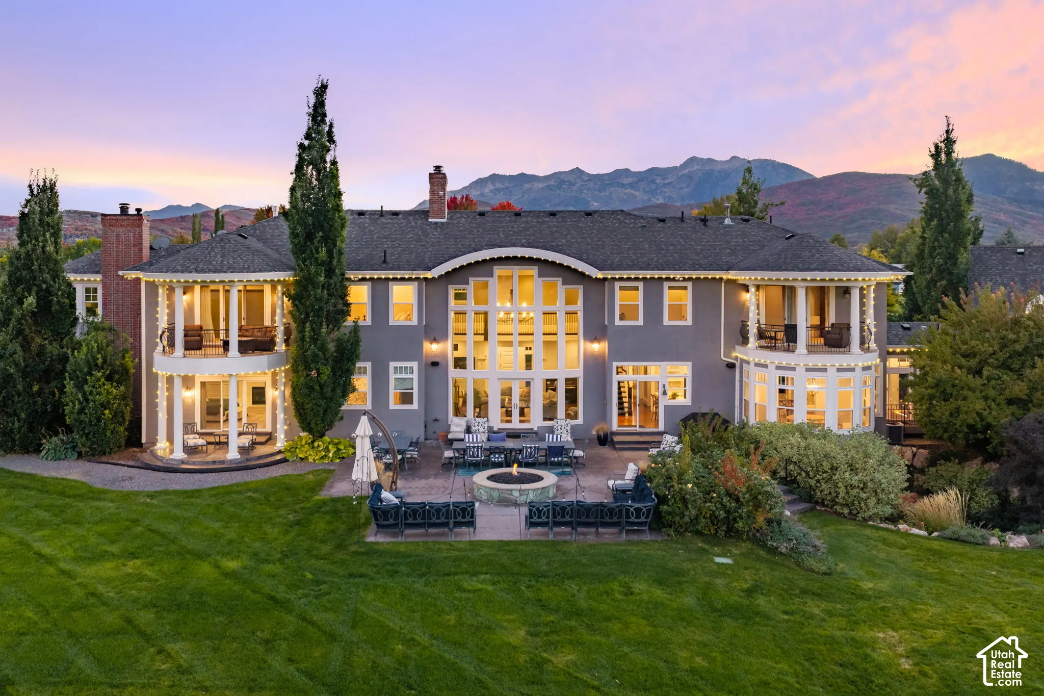 Expansive residence featuring two-story multi-pane windows, dual covered balconies with circular architectural detail, stone-surround fire pit, and professional landscaping against a mountain backdrop