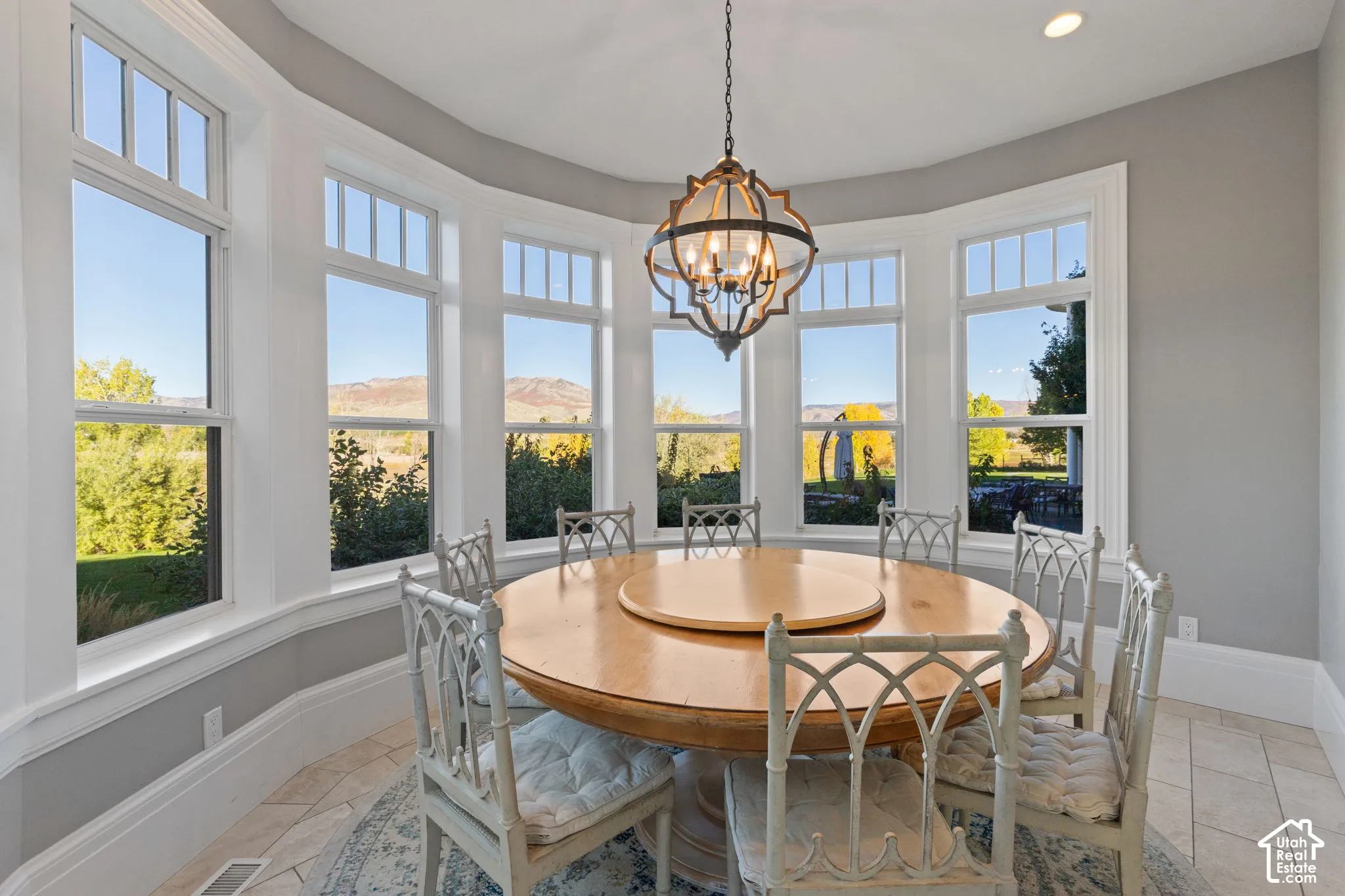 Elegant dining room showcasing a curved bay of expansive multi-pane windows framing panoramic landscape vistas, hardwood flooring, recessed ceiling detail, a coordinating geometric pendant light fixture, and ample natural illumination throughout the space