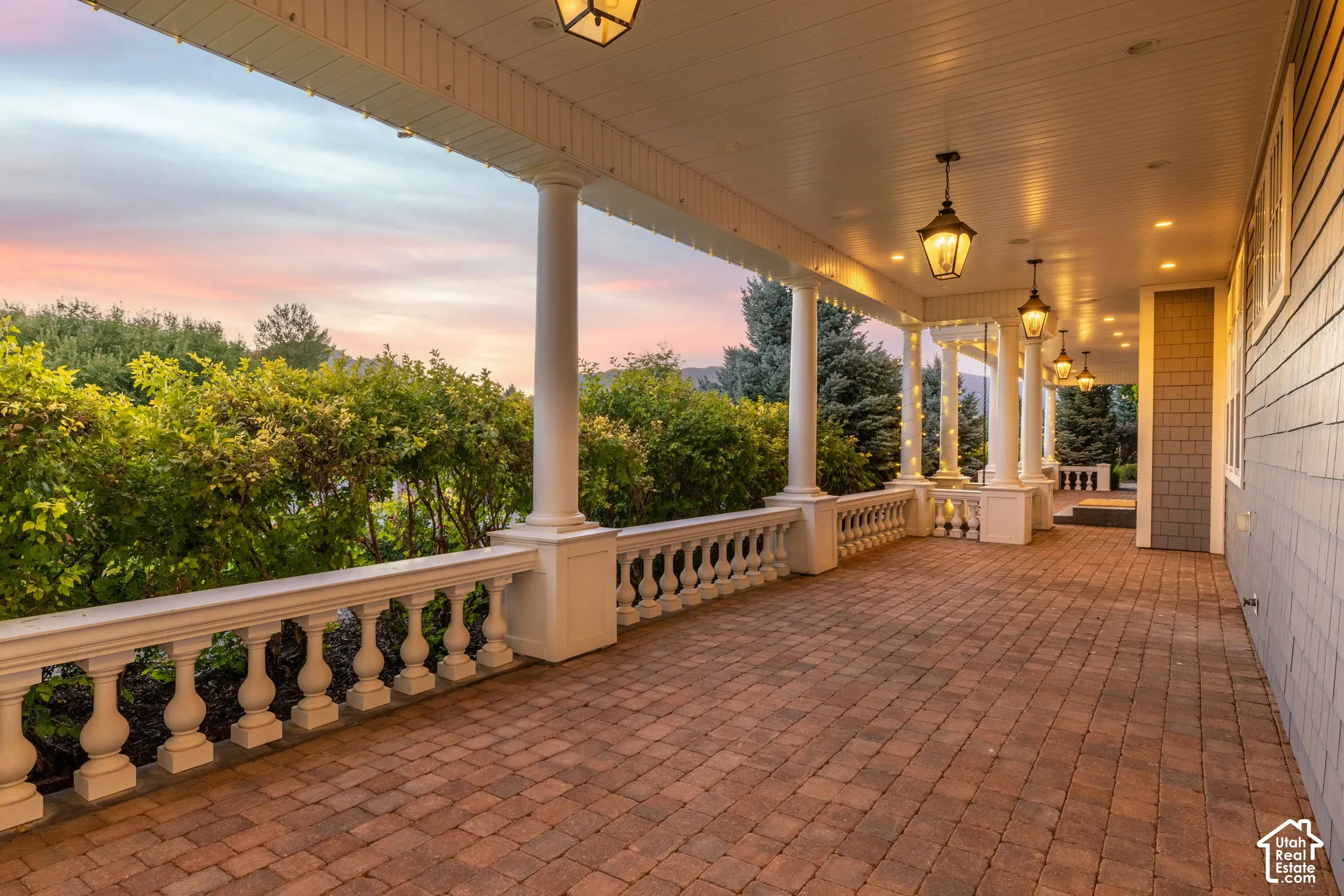 Covered veranda featuring decorative balustrade railings, architectural columns, brick-paver flooring, and hanging lantern-style fixtures